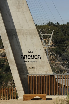 A large concrete structure labeled '516 Arouca Ponte Suspensa' appears prominently. The structure is part of a suspension bridge, with steel cables extending into the background. Wooden railings and a bench are at the base of the structure, and green hills are visible in the distance.