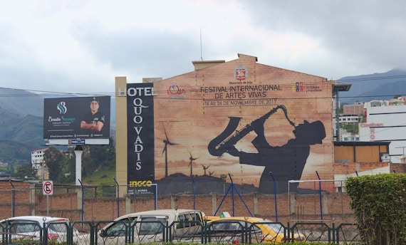 A large mural on the side of a building depicts a silhouette of a person playing a saxophone, promoting a festival. The mural is set against a backdrop of wind turbines. Nearby, there's a billboard featuring a person advertising services, and a hotel sign with 'Quo Vadis'. The scene includes parked vehicles and mountains in the distance under a cloudy sky.