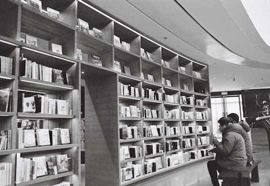 A large bookshelf spans across the wall, filled with a variety of books organized neatly. Two people are seated in front of the shelves, reading intently. The setting appears to be a library or bookstore with a calm and quiet atmosphere. The image is in black and white, adding a classic and timeless feel.