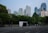 A bustling urban landscape with modern skyscrapers towering over a lush, green park. Several people are casually walking or jogging along a wide road in the foreground, creating a lively atmosphere amidst the tranquil greenery. The glass and steel structures of the buildings reflect the clear blue sky, adding a sense of openness to the scene.