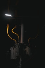 A dimly lit scene features a streetlight illuminating part of a utility pole with several cylindrical transformers. The cables are prominently highlighted in yellow against the dark background.