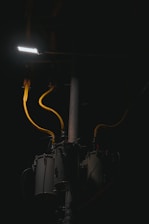 A dimly lit scene features a streetlight illuminating part of a utility pole with several cylindrical transformers. The cables are prominently highlighted in yellow against the dark background.