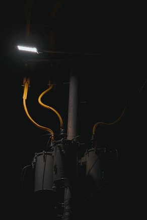 A dimly lit scene features a streetlight illuminating part of a utility pole with several cylindrical transformers. The cables are prominently highlighted in yellow against the dark background.