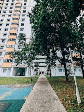 A tall residential building with a modern design stands next to a walkway lined with grass and trees. The building features multiple stories with balconies, large windows, and a white and orange color scheme. Bicycles are parked under the building's overhang, and a lamp post is visible in the foreground.