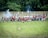 A historical reenactment scene with individuals dressed in period military uniforms, some in red coats and others in white attire. They are engaged in a simulated battle, with smoke visible from a cannon or musket fire. The setting is an open grassy field with dense green trees in the background.