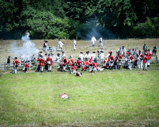 A historical reenactment scene with individuals dressed in period military uniforms, some in red coats and others in white attire. They are engaged in a simulated battle, with smoke visible from a cannon or musket fire. The setting is an open grassy field with dense green trees in the background.