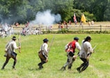 A historical reenactment of a battle scene with colonial soldiers dressed in period uniforms engaged in combat on a grassy field. Smoke rises from musket fire as soldiers advance and take cover. The backdrop includes a wooden fence and a mixed forest area. Flags are visible, adding to the historic ambiance.