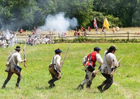 A historical reenactment of a battle scene with colonial soldiers dressed in period uniforms engaged in combat on a grassy field. Smoke rises from musket fire as soldiers advance and take cover. The backdrop includes a wooden fence and a mixed forest area. Flags are visible, adding to the historic ambiance.