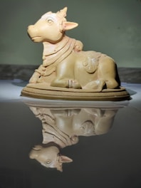A small, detailed sculpture of a cow adorned with intricate carvings is placed on a reflective surface, creating a mirrored image below it. The cow is shown in a sitting position with ornate designs on its body and wearing a necklace.