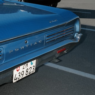 A vintage blue Pontiac GTO, featuring a Swiss license plate, is parked in a lot. The rear section of the car is visible, showing the brand logo and tail lights.