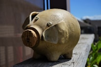 A ceramic piggy bank with spiral patterns is placed on a wooden surface. The bank is crafted in an earthy tone, shaped with minimal features including two small eyes and a cork stopper as the snout. Sunlight casts shadows, suggesting an outdoor setting against a backdrop of a building and a clear blue sky.