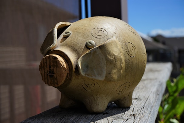 A ceramic piggy bank with spiral patterns is placed on a wooden surface. The bank is crafted in an earthy tone, shaped with minimal features including two small eyes and a cork stopper as the snout. Sunlight casts shadows, suggesting an outdoor setting against a backdrop of a building and a clear blue sky.