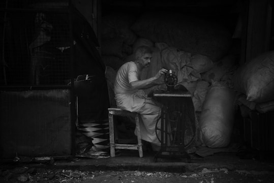 An older man is sitting on a wooden chair operating a manual sewing machine. He is surrounded by large bags filled with materials in a dimly lit room. The scene appears to be inside a workshop or storage area.