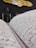 A close-up view of an open book with Arabic script, likely a religious text. In the background, there is a decorative plaque with calligraphy and ornate details, possibly hanging on a wall. The setting appears to be indoors with dim lighting, featuring a chair and a stand with papers.