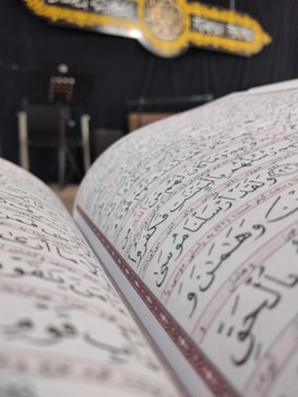 A close-up view of an open book with Arabic script, likely a religious text. In the background, there is a decorative plaque with calligraphy and ornate details, possibly hanging on a wall. The setting appears to be indoors with dim lighting, featuring a chair and a stand with papers.