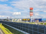 A large retail complex is visible with prominent signs for several businesses including IKEA, JULA, and TK Maxx. The stores are positioned behind a road with a tall metal barrier. There is a bright blue sky with scattered clouds, and green grass lines the sides of the road.