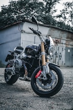 A sleek motorcycle with a shiny chrome finish and gold fork tubes is parked on a gravel surface. The motorcycle features a prominent front headlight, black body, and red brake calipers. It is positioned in front of a rustic building with overgrown foliage and a weathered metal door in the background.