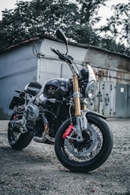 A sleek motorcycle with a shiny chrome finish and gold fork tubes is parked on a gravel surface. The motorcycle features a prominent front headlight, black body, and red brake calipers. It is positioned in front of a rustic building with overgrown foliage and a weathered metal door in the background.