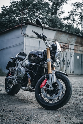 A sleek motorcycle with a shiny chrome finish and gold fork tubes is parked on a gravel surface. The motorcycle features a prominent front headlight, black body, and red brake calipers. It is positioned in front of a rustic building with overgrown foliage and a weathered metal door in the background.