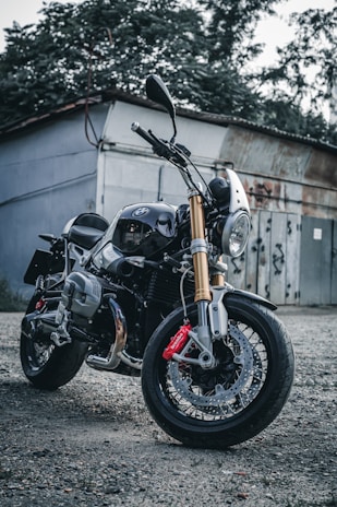 A sleek motorcycle with a shiny chrome finish and gold fork tubes is parked on a gravel surface. The motorcycle features a prominent front headlight, black body, and red brake calipers. It is positioned in front of a rustic building with overgrown foliage and a weathered metal door in the background.