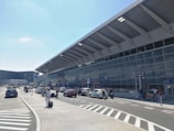 The exterior of a modern airport terminal with a large glass facade and a slanted roof. Cars, taxis, and people with luggage are seen in the drop-off and pick-up area along the road. There are visible signs indicating different zones and parking areas, as well as several people walking or standing with their suitcases. The sky is clear with minimal clouds.