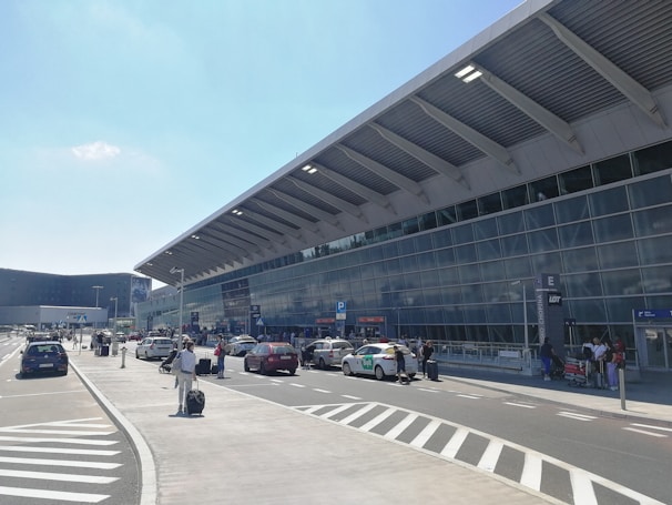 The exterior of a modern airport terminal with a large glass facade and a slanted roof. Cars, taxis, and people with luggage are seen in the drop-off and pick-up area along the road. There are visible signs indicating different zones and parking areas, as well as several people walking or standing with their suitcases. The sky is clear with minimal clouds.