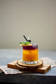 A vibrant cocktail in a transparent glass, showcasing a gradient of deep purple at the top blending into a bright orange-yellow below. Garnished with a slice of citrus fruit and fresh herbs, the drink is set on circular wooden coasters over a newspaper on a gray surface.