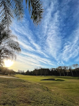 A picturesque golf course with well-maintained green grass and sand bunkers is seen under a vibrant sky filled with scattered clouds. The sun is setting or rising, casting a warm glow over the landscape. Tall palm trees frame the image, adding a tropical feel to the scene. In the distance, leafless trees border the horizon.