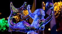 An intricately carved ice sculpture of a mermaid is illuminated with colorful lights, creating a radiant glow. The mermaid is holding a spherical object, possibly representing a ball, in one hand. The background features festive, blurred bokeh lights in shades of yellow and green, enhancing the celebratory atmosphere.