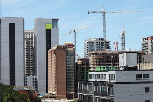 High-rise buildings and construction cranes dominate the urban landscape. A combination of modern skyscrapers and mid-rise buildings, with a construction site featuring cranes. Some buildings have signage, indicating institutional use.