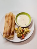 A white plate contains two crispy golden brown rolled dosas, a green-rimmed white bowl filled with creamy white sauce garnished with black pepper, and a side of roasted cauliflower and red bell pepper pieces.