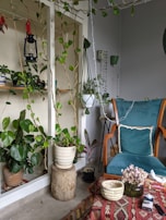 A cozy indoor space filled with greenery, featuring a variety of potted plants on shelves and hanging from the ceiling. A vintage lantern and a dreamcatcher hang as decorative pieces. A comfortable armchair with a teal cushion is positioned near a table covered with a patterned cloth, on which rests a woven basket, a vase with dried flowers, and a box of tissues.