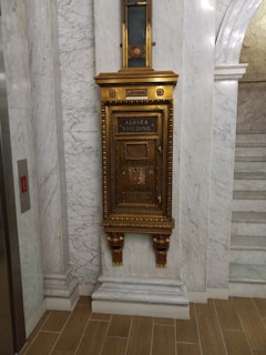 A vintage, ornate mail chute is mounted on a marble wall. The chute is elaborately decorated with gold trimming and features the text 'Alaska Building' and 'U.S. Mail'. The surrounding area includes marble paneling and a wooden floor.