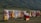 A colorful display of traditional clothing hanging on racks is set against a lush green hillside. Garments in various vibrant colors such as red, yellow, blue, and pink are shown. A small stall with a red cloth on top offers various items, possibly headwear or accessories.