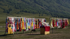 A colorful display of traditional clothing hanging on racks is set against a lush green hillside. Garments in various vibrant colors such as red, yellow, blue, and pink are shown. A small stall with a red cloth on top offers various items, possibly headwear or accessories.