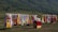 A colorful display of traditional clothing hanging on racks is set against a lush green hillside. Garments in various vibrant colors such as red, yellow, blue, and pink are shown. A small stall with a red cloth on top offers various items, possibly headwear or accessories.