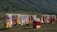 A colorful display of traditional clothing hanging on racks is set against a lush green hillside. Garments in various vibrant colors such as red, yellow, blue, and pink are shown. A small stall with a red cloth on top offers various items, possibly headwear or accessories.