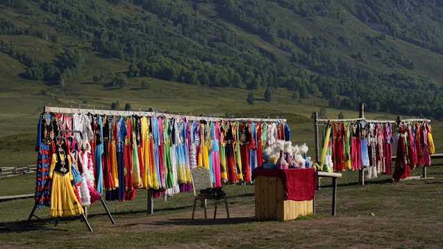A colorful display of traditional clothing hanging on racks is set against a lush green hillside. Garments in various vibrant colors such as red, yellow, blue, and pink are shown. A small stall with a red cloth on top offers various items, possibly headwear or accessories.