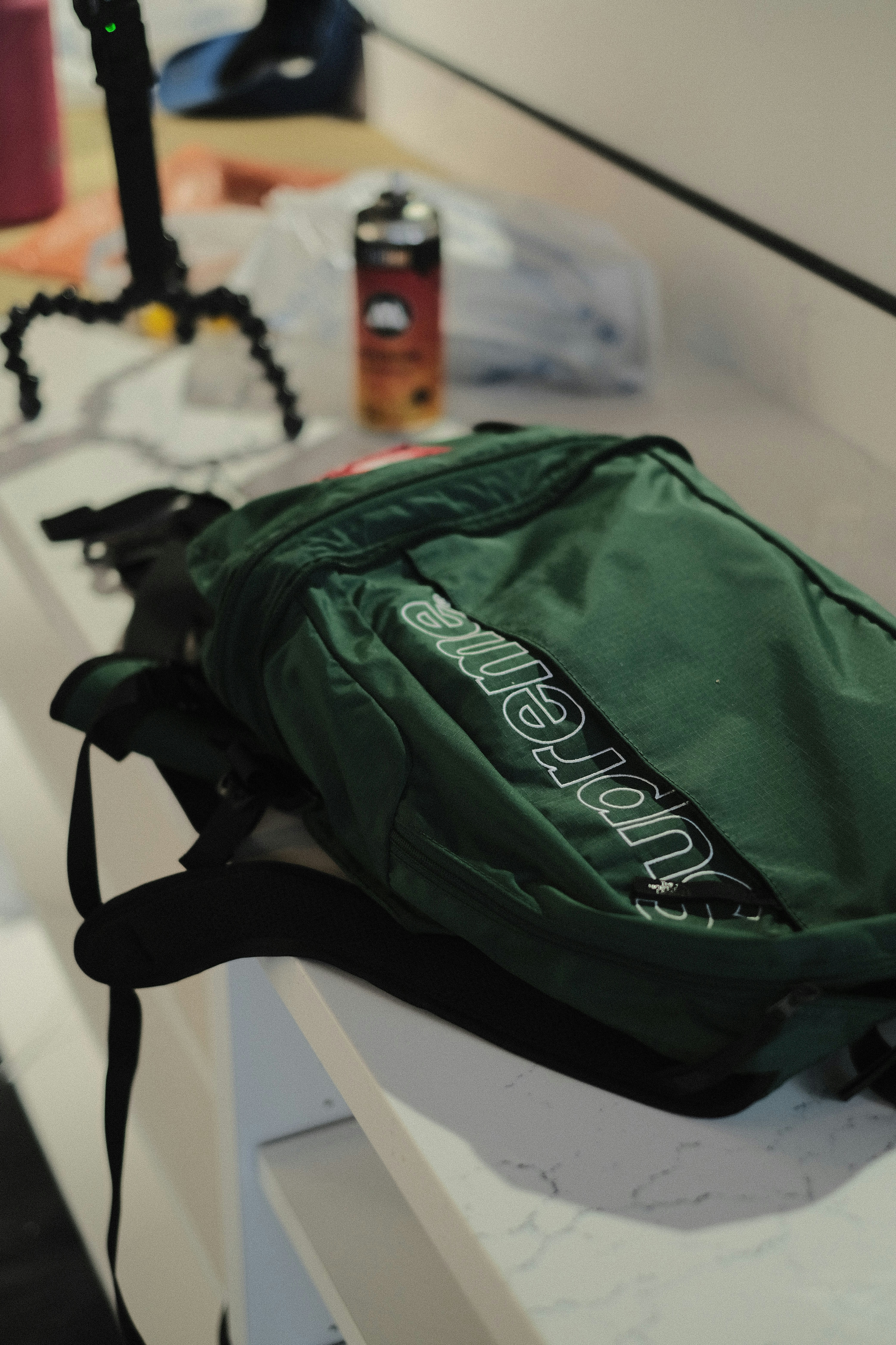 A green backpack with a prominent logo is placed on a white counter. Nearby, there is a drink can and a flexible tripod. The scene suggests a casual, everyday setting with accessories on a countertop.
