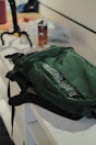 A green backpack with a prominent logo is placed on a white counter. Nearby, there is a drink can and a flexible tripod. The scene suggests a casual, everyday setting with accessories on a countertop.