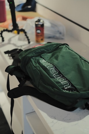 A green backpack with a prominent logo is placed on a white counter. Nearby, there is a drink can and a flexible tripod. The scene suggests a casual, everyday setting with accessories on a countertop.