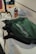 A green backpack with a prominent logo is placed on a white counter. Nearby, there is a drink can and a flexible tripod. The scene suggests a casual, everyday setting with accessories on a countertop.