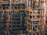 An intricate network of industrial metal structures and machinery with several tall cylindrical towers interconnected by yellow safety railings. The complex design and metallic components indicate a large-scale industrial facility, possibly a refinery or power plant.