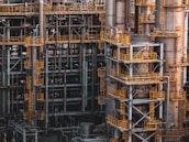 An intricate network of industrial metal structures and machinery with several tall cylindrical towers interconnected by yellow safety railings. The complex design and metallic components indicate a large-scale industrial facility, possibly a refinery or power plant.