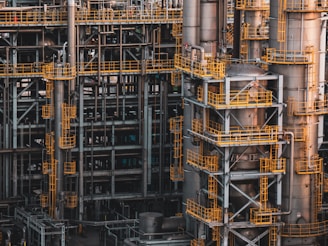 An intricate network of industrial metal structures and machinery with several tall cylindrical towers interconnected by yellow safety railings. The complex design and metallic components indicate a large-scale industrial facility, possibly a refinery or power plant.