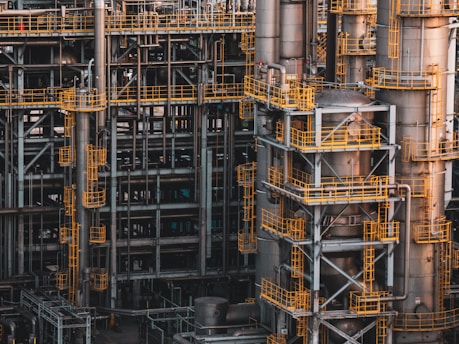 An intricate network of industrial metal structures and machinery with several tall cylindrical towers interconnected by yellow safety railings. The complex design and metallic components indicate a large-scale industrial facility, possibly a refinery or power plant.