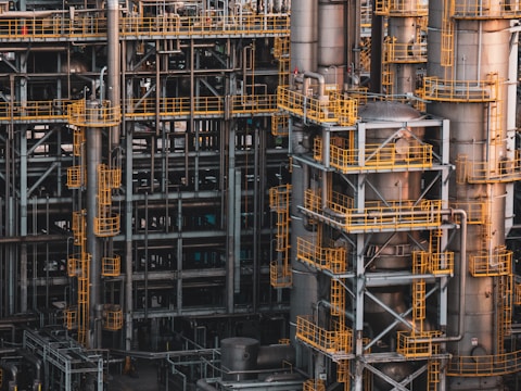 An intricate network of industrial metal structures and machinery with several tall cylindrical towers interconnected by yellow safety railings. The complex design and metallic components indicate a large-scale industrial facility, possibly a refinery or power plant.