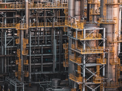An intricate network of industrial metal structures and machinery with several tall cylindrical towers interconnected by yellow safety railings. The complex design and metallic components indicate a large-scale industrial facility, possibly a refinery or power plant.
