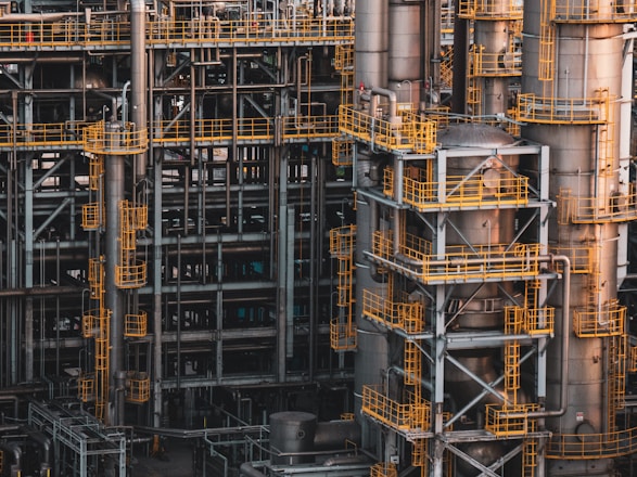 An intricate network of industrial metal structures and machinery with several tall cylindrical towers interconnected by yellow safety railings. The complex design and metallic components indicate a large-scale industrial facility, possibly a refinery or power plant.