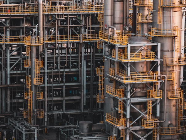 An intricate network of industrial metal structures and machinery with several tall cylindrical towers interconnected by yellow safety railings. The complex design and metallic components indicate a large-scale industrial facility, possibly a refinery or power plant.